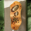 A top-down view of a rustic, irregularly shaped wooden coffee table with natural burl wood grain, featuring two carved stone bowls and a bouquet of flowers, set on a textured beige rug between green velvet couches.