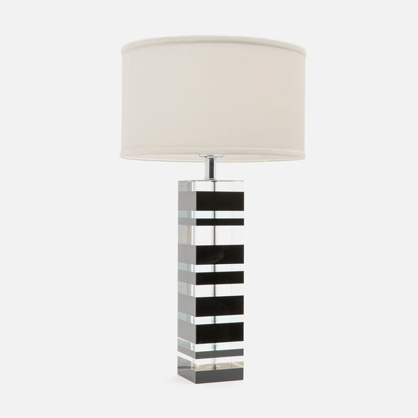 A modern table lamp with a cylindrical off-white fabric drum shade and a geometric column base made of alternating black and clear acrylic or glass rectangular panels, set against a plain light gray background.