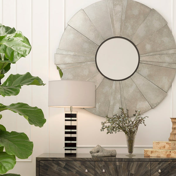 A large circular wall mirror with a sunburst design and distressed silver-gray metallic finish, mounted on a white paneled wall above a console table with a lamp, plant, and decorative items.