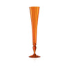 An amber orange translucent glass champagne flute with a flared rim and a turned stem, standing on a reflective white surface against a white background.