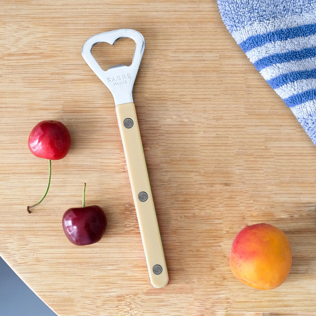 A bottle opener with a cream-colored handle and a silver metal head featuring a heart-shaped cutout, placed on a light wood cutting board next to two red cherries and a peach, with a blue and white striped towel in the corner.