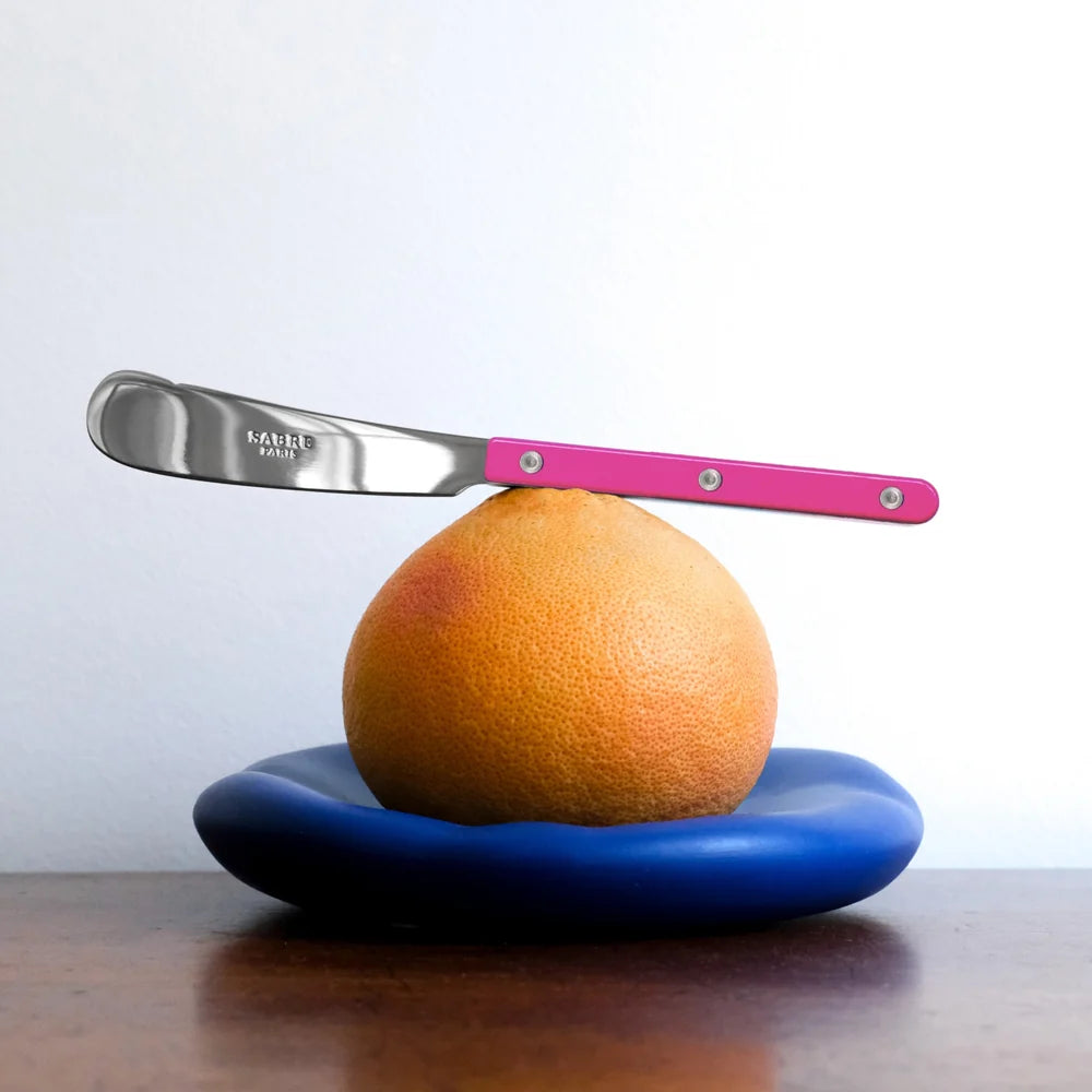 Knife with pink handle balancing on an orange on a blue plate against a white background