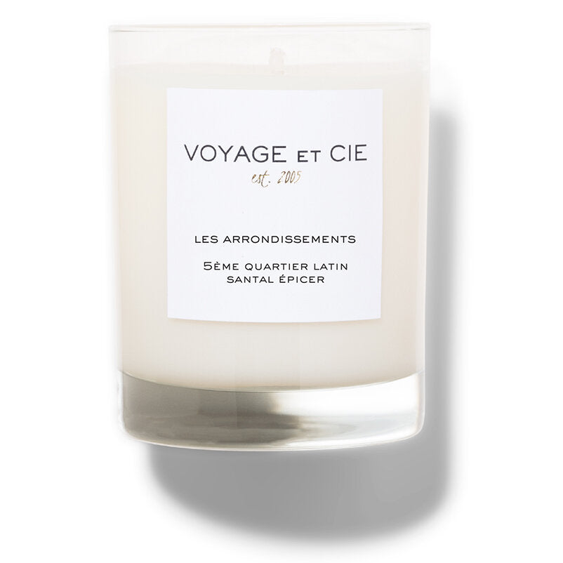 A beige candle with a white label that reads 'Voyage et Cie Les Arrondissements Seme Quartier Latin Santal Epicer'.