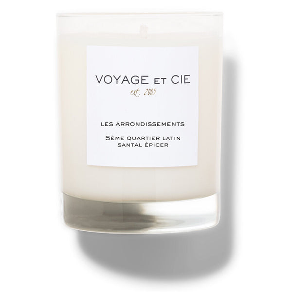 A beige candle with a white label that reads 'Voyage et Cie Les Arrondissements Seme Quartier Latin Santal Epicer'.