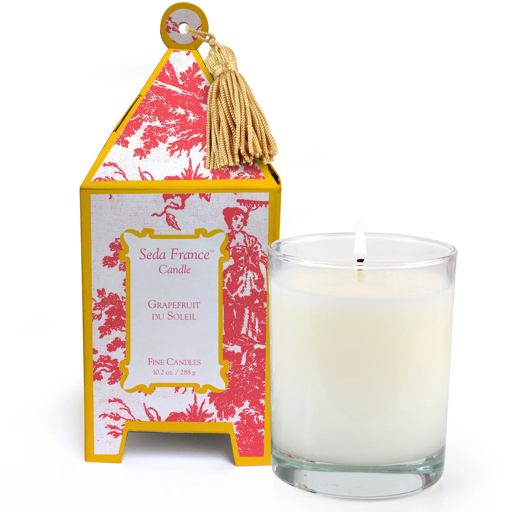 Candle in glass container next to yellow and pink patterned pagoda box on white background
