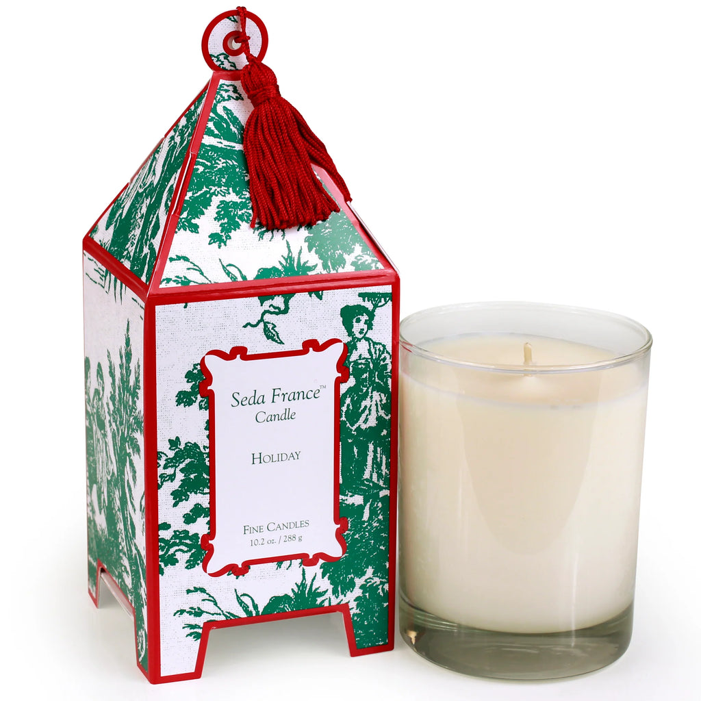 A Seda France 'Holiday' scented candle shown next to its decorative packaging. The packaging features a green and white toile pattern with a red tassel on top, and the candle itself is in a clear glass jar filled with cream-colored wax.