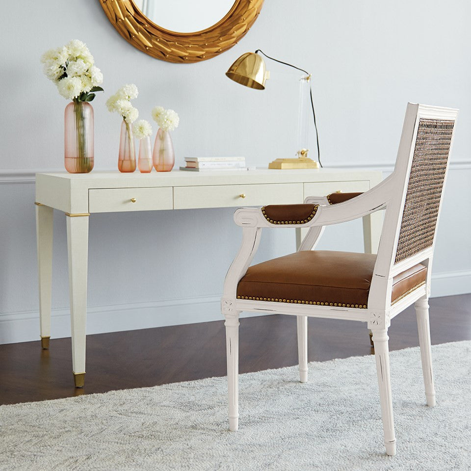 A white upholstered armchair with a brown cushion and woven cane back is placed in front of a matching white desk with gold-tone hardware in a bright room. A gold floor lamp, pink glass vases with white flowers, and a round gold mirror are also visible.