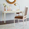 A white upholstered armchair with a brown cushion and woven cane back is placed in front of a matching white desk with gold-tone hardware in a bright room. A gold floor lamp, pink glass vases with white flowers, and a round gold mirror are also visible.