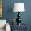 A navy blue bedside table with gold trim and brass legs stands against a dark blue wall, styled with a matching navy table lamp, a white drum shade, and a small gold decorative object. Part of a white armchair is visible on the left.