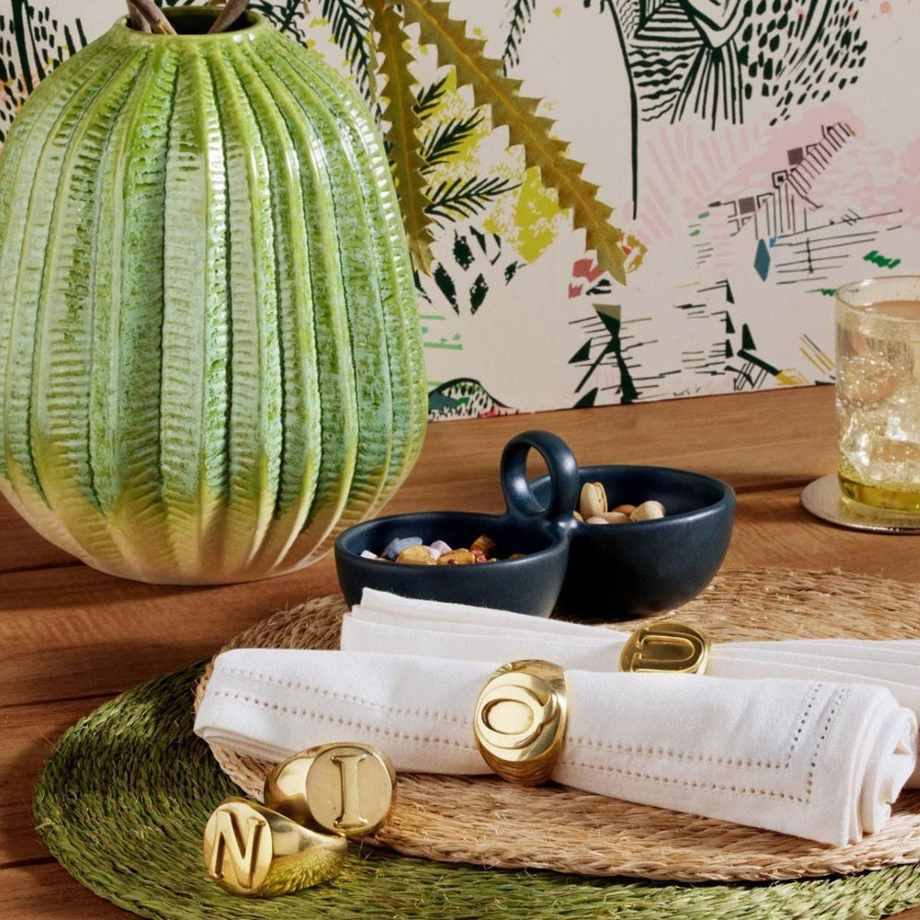 Gold-finished brass napkin rings with a customizable design, displayed on a woven mat alongside a green decorative object, with a glass of liquid and a bowl of nuts in the background.
