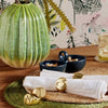 Gold-finished brass napkin rings with a customizable design, displayed on a woven mat alongside a green decorative object, with a glass of liquid and a bowl of nuts in the background.